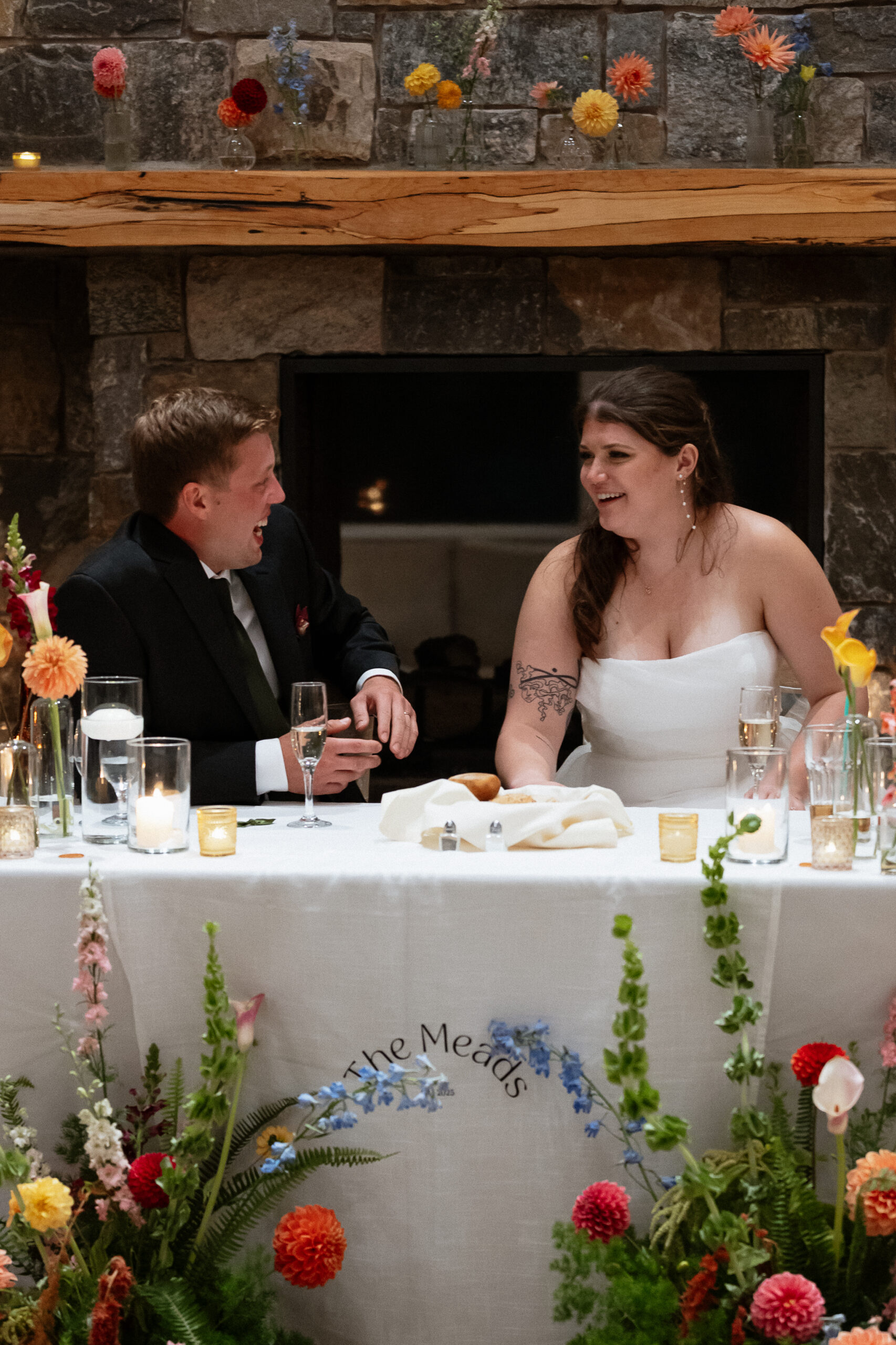 Wedding Photographer VT | Sincerely Liz Photography Documentary and Editorial Wedding Photography | A couple in wedding attire sits at a flower-adorned table, smiling warmly at each other. The scene conveys joy and celebration, set against a rustic stone backdrop.