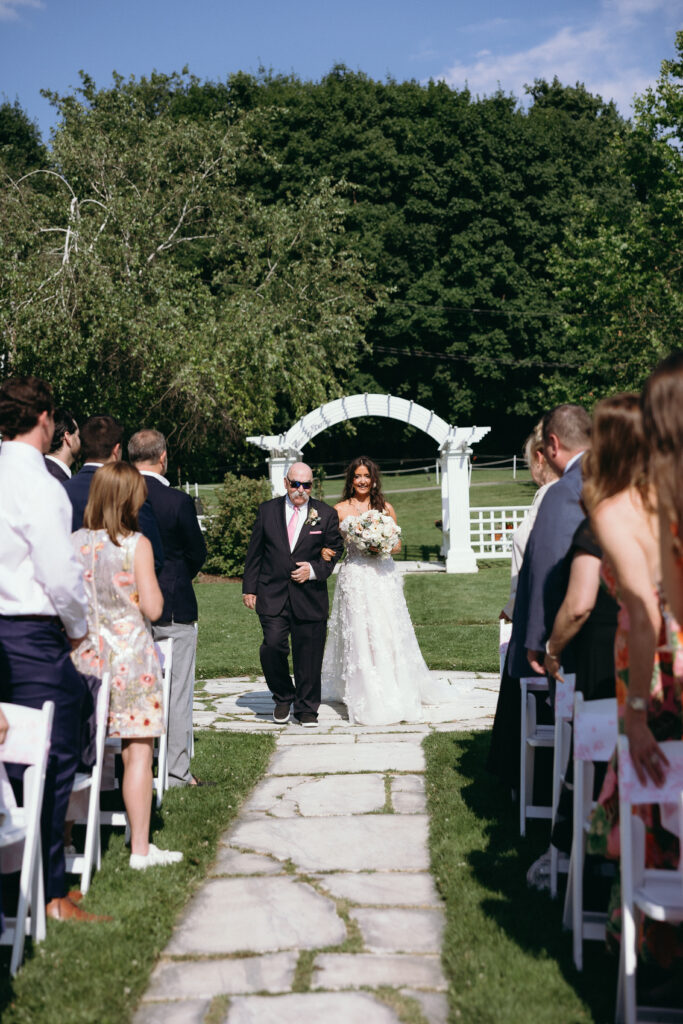 Bride walking down a stone aisle with her father as guests look on during an outdoor ceremony, a heartfelt moment from an intimate Vermont wedding.