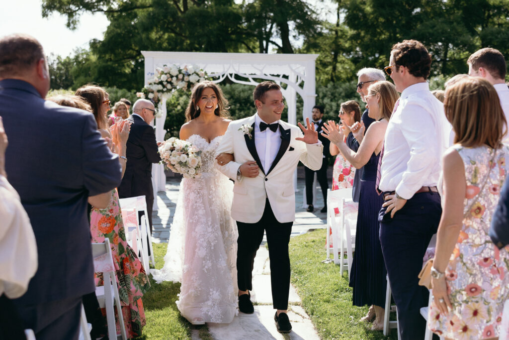 Wedding Photographer in Vermont | Sincerely Liz Photography Documentary and Editorial Wedding Photography | Bride and groom joyfully walk down the aisle outdoors, surrounded by clapping guests. The bride holds a bouquet, and they both smile brightly.