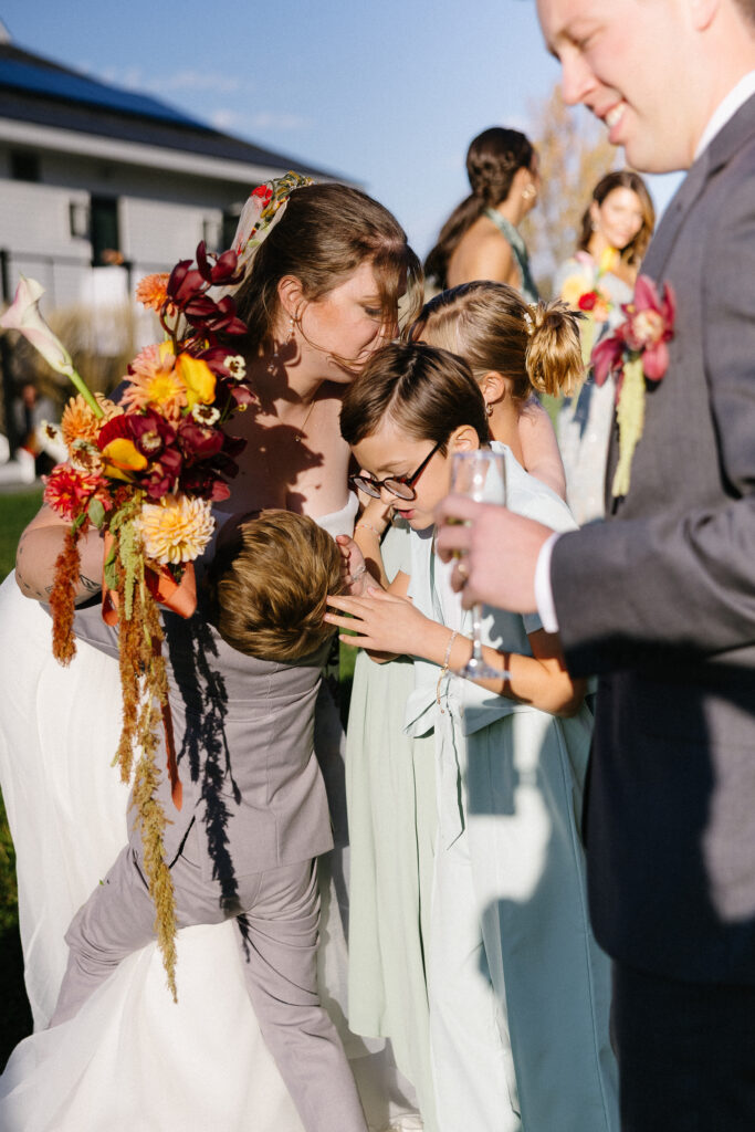 Wedding Photographer VT | Sincerely Liz Photography Documentary and Editorial Wedding Photography | A woman in a strapless dress holds a vibrant bouquet as she embraces three smiling children. A man in a suit stands nearby, holding a drink. The atmosphere is joyful and warm under clear skies.