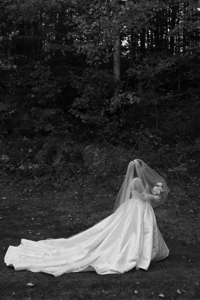 Wedding Photographer in Vermont | Sincerely Liz Photography Documentary and Editorial Wedding Photography | A bride in a flowing gown and veil stands in a wooded area, her back turned. Holding a bouquet, she exudes elegance against the dark, leafy backdrop.