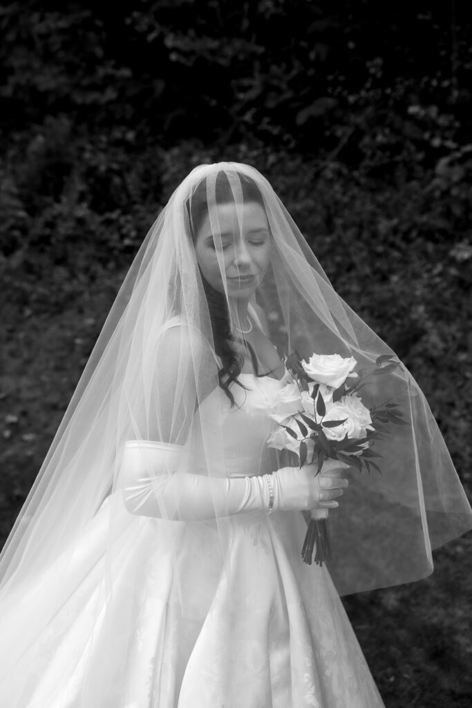 Wedding Photographers in Vermont | Sincerely Liz Photography Documentary and Editorial Wedding Photography | A bride in a flowing gown and veil holds a bouquet of roses against a lush, leafy background. Her eyes are closed, conveying a serene emotion. Black and white.