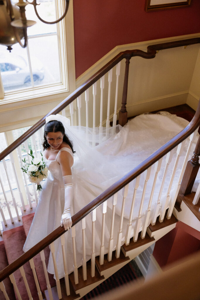 Wedding Photographers in Vermont | Sincerely Liz Photography Documentary and Editorial Wedding Photography | A bride in a flowing white dress and veil descends a wooden staircase, holding a bouquet. The setting feels elegant and joyous.