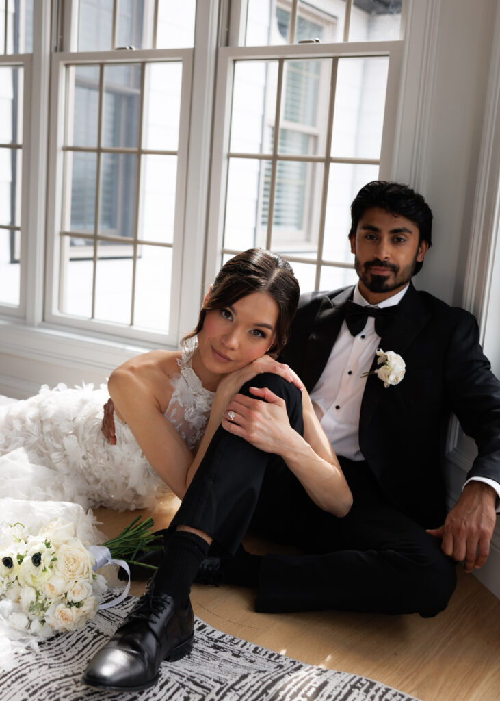 The newlyweds sit together on the floor by tall windows in soft natural light, an intimate portrait from a wedding in Vermont.