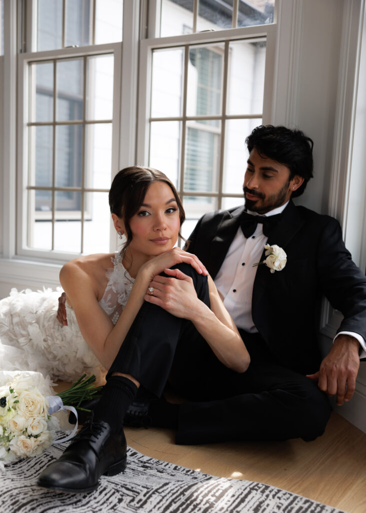 Bride and groom sit together on the floor beside tall windows during a relaxed indoor portrait session, showcasing the candid connection often prioritized by wedding photographers in Massachusetts.