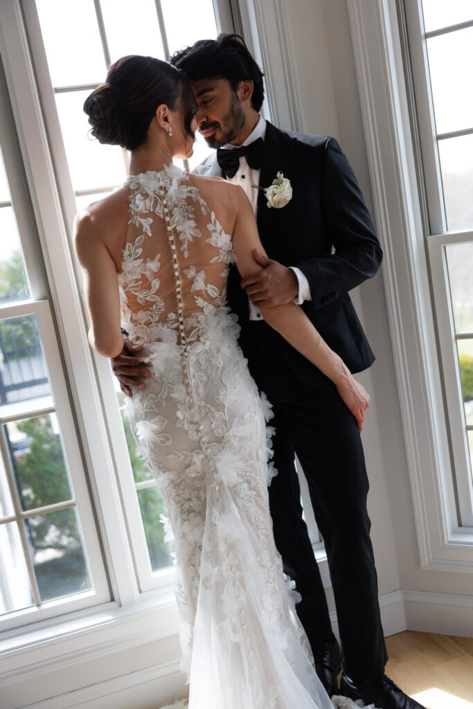 Bride and groom stand beside tall windows in an intimate portrait highlighting the texture of her floral gown and the soft natural light, photographed in the refined style often associated with Massachusetts wedding photographers.