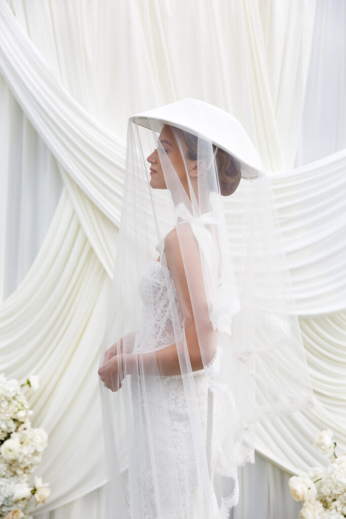 Bride in a sculptural veil and wide-brim hat stands against softly draped fabric in an editorial bridal portrait, reflecting the refined style often associated with luxury wedding photography VT.