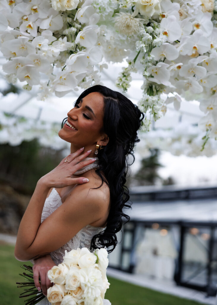 Bride smiles beneath cascading white florals while holding a bouquet of roses during an outdoor portrait session, a timeless approach to fine-art Vermont wedding photographer imagery.