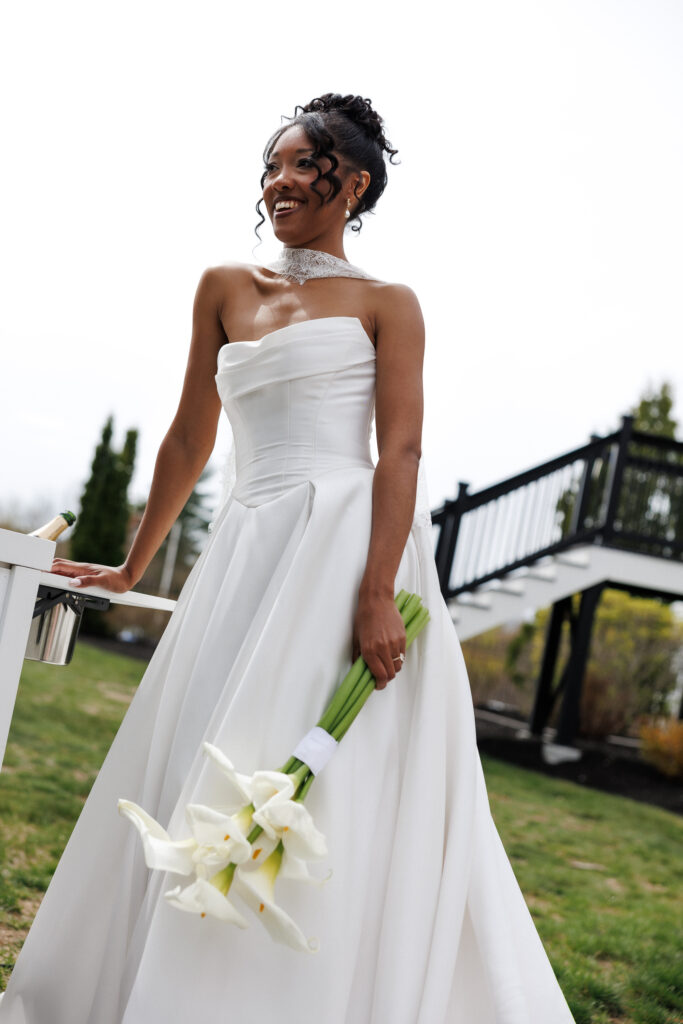 A bride stands outdoors holding white calla lilies while looking off to the side in soft daylight, a refined portrait for elegant Vermont wedding venues.
