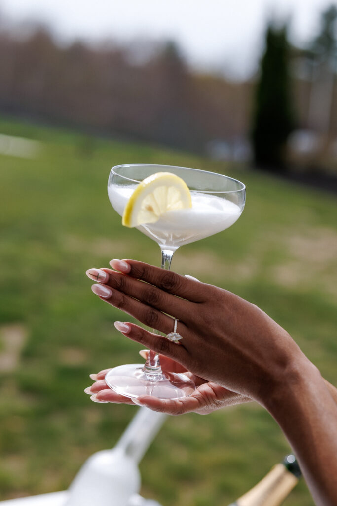 Bride holds a coupe glass garnished with lemon during an outdoor cocktail hour portrait, adding a playful editorial moment to this romantic wedding photography Massachusetts gallery.