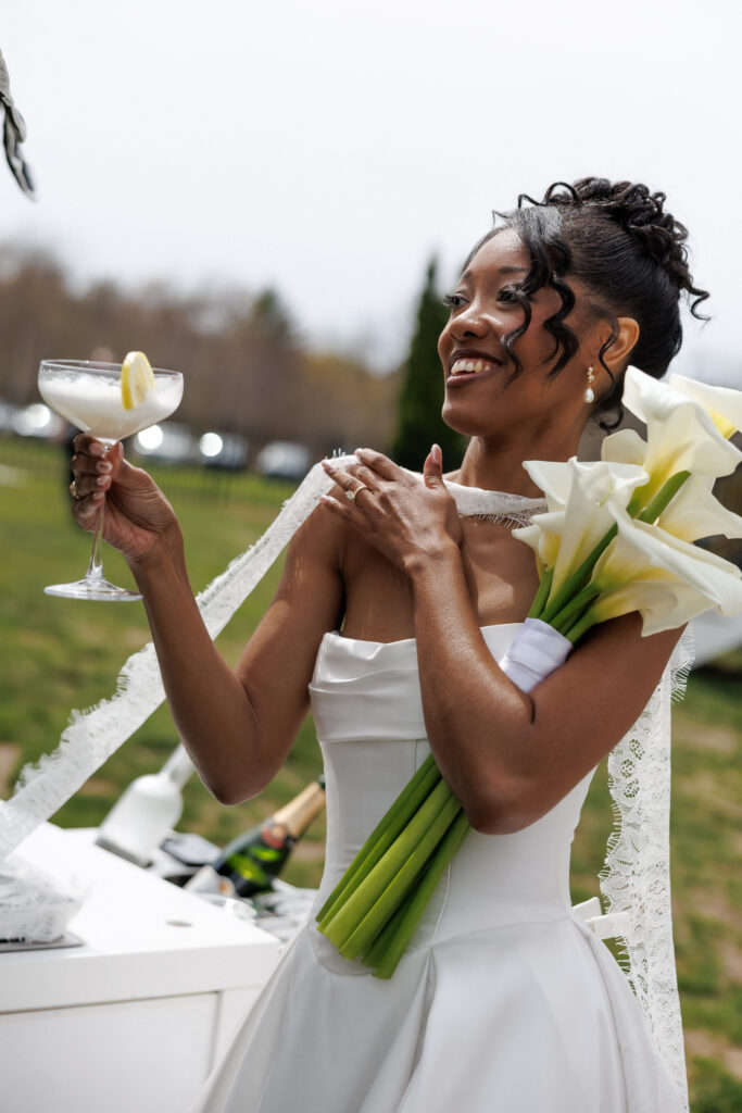 Bride smiles outdoors while holding white calla lilies and a signature cocktail during a playful portrait moment inspired by Singing in the Rain wedding details and artistic wedding photographers in Massachusetts.