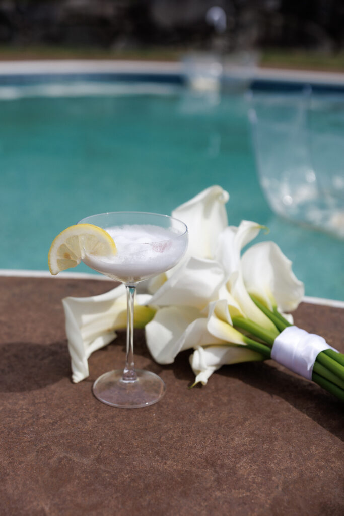 Signature cocktail with citrus garnish and white calla lilies rests beside a poolside setting during wedding preparations, a detail-focused moment captured through artistic Vermont wedding photography.