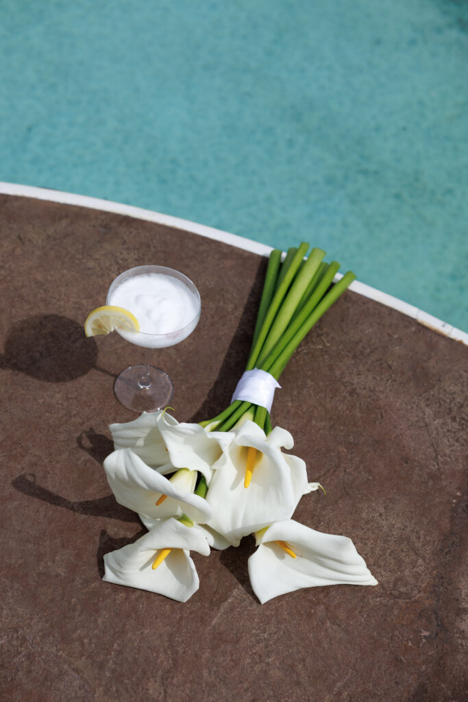 Flat lay of white calla lilies and a citrus cocktail beside a turquoise pool photographed from above, reflecting the intentional composition and true-to-tone style many Massachusetts wedding photographers are known for.