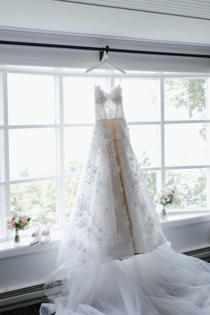 White floral lace wedding gown hanging beside a bright window with small flower arrangements