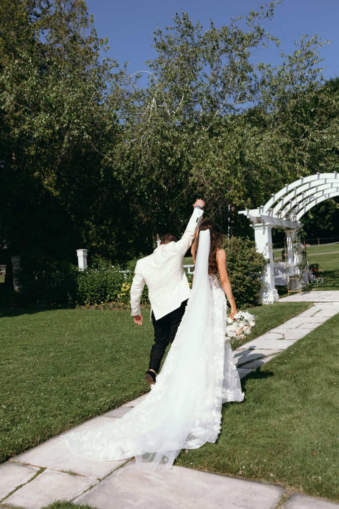 Newlyweds walking hand in hand along a stone pathway beside a white garden arch at Basin Harbor